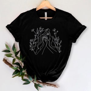 May include: Black t-shirt with a white line drawing of a woman's face and flowing hair, framed by floral motifs. The minimalist design provides a strong contrast against the dark background. The t-shirt is displayed on a wooden hanger.