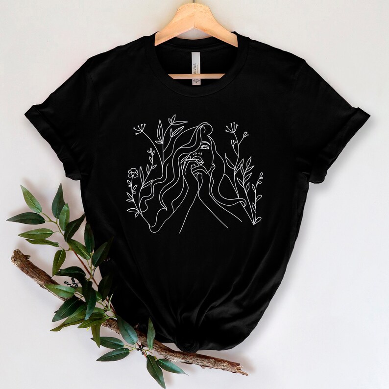 May include: Black t-shirt with a white line drawing of a woman's face and flowing hair, framed by floral motifs. The minimalist design provides a strong contrast against the dark background. The t-shirt is displayed on a wooden hanger.