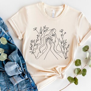 May include: A cream-coloured t-shirt with a black line drawing of a woman's face and flowing hair, framed by floral details. The design is centrally placed on the shirt, which has short sleeves and a crew neck. A denim jacket is next to the shirt.