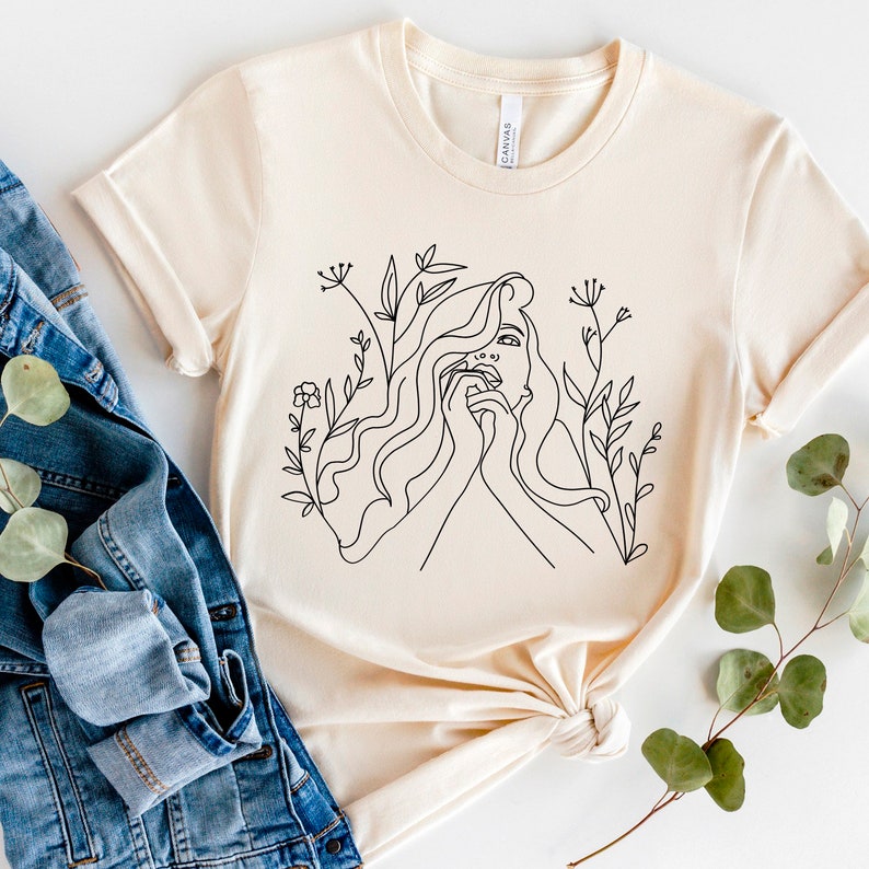 May include: A cream-coloured t-shirt with a black line drawing of a woman's face and flowing hair, framed by floral details. The design is centrally placed on the shirt, which has short sleeves and a crew neck. A denim jacket is next to the shirt.