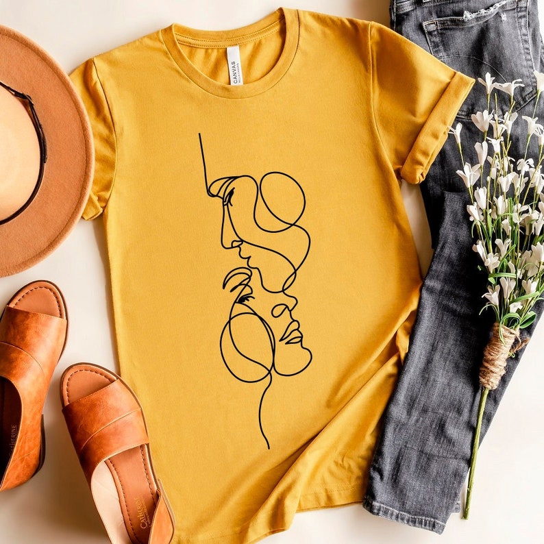 May include: A mustard yellow t-shirt featuring a black line drawing of two faces intertwined. The faces are stylised and abstract.