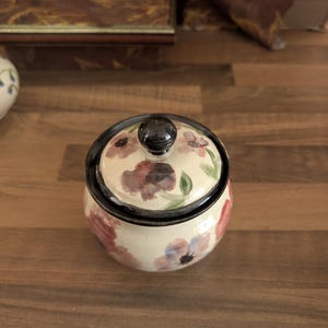 May include: A small, round ceramic jar with a matching lid, featuring a hand-painted floral pattern in shades of pink, purple, and green on a cream-coloured background. The lid has a dark brown knob and a dark brown rim, matching the jar's base.