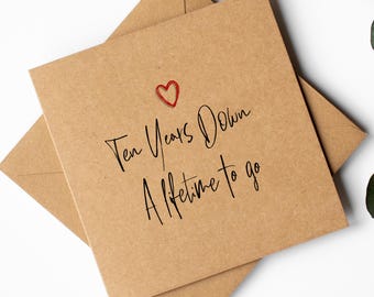 May include: A kraft paper card with the text "Ten Years Down, A lifetime to go" in black script, topped with a red heart. The card sits on a kraft paper envelope. Ideal for an anniversary.