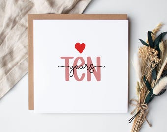May include: A square greeting card with a brown border and a white interior. The card features the text "10 years" in a script font, with a red heart above the number 10. A bouquet of dried flowers is to the right.