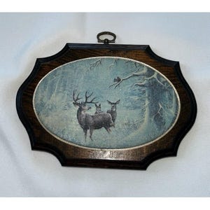 May include: An oval wall plaque featuring a winter forest scene with three deer, including one buck with antlers. The artwork is set within a dark brown, scalloped wooden frame with a metallic silver border and a small metal hanging loop at the top.