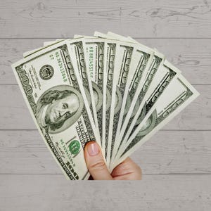 May include: A hand holding a fan of ten US dollar bills. The bills are fanned out in a semi-circle, with the top bill showing the portrait of Benjamin Franklin and the text "FEDERAL RESERVE NOTE" and "KB 881455155".