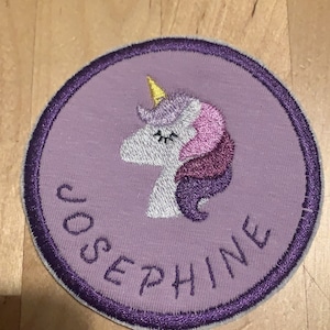 Könnte beinhalten: Lilafarbener gestickter Aufnäher mit einem weißen Einhorn mit einem gelben Horn und einer rosa und lila Mähne. Der Aufnäher trägt die Aufschrift "Josephine" in einer lilafarbenen Kursivschrift.