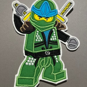 Könnte beinhalten: Grüne und blaue gestickte Applikation einer Lego Minifigur Ninja, die zwei Schwerter hält. Der Ninja trägt einen schwarzen Gürtel und ein blaues Stirnband.