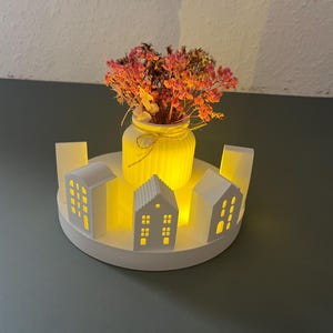 Könnte beinhalten: Eine weiße Keramik-Tischdekoration mit einem runden Sockel und fünf kleinen Häusern mit ausgeschnittenen Fenstern. Eine Glasvase mit getrockneten Blumen steht in der Mitte des Sockels.