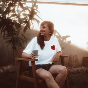 Peut inclure: Une femme portant un t-shirt blanc avec un motif de fleur rouge est assise sur une chaise en bois dans un jardin. Elle tient une tasse en céramique verte et sourit.