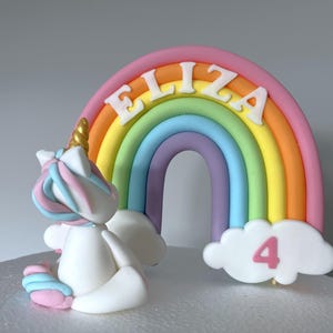 A cake topper with a white unicorn and a rainbow-colored arch with the name "ELIZA" written on it.