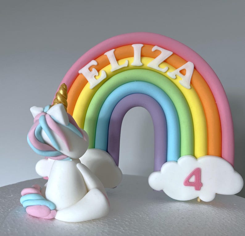A cake topper with a white unicorn and a rainbow-colored arch with the name "ELIZA" written on it.