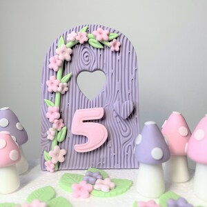 May include: Purple and pink fondant cake topper featuring a wooden door design, a heart-shaped window, and the number 5. The door is decorated with a vine of pink flowers and green leaves. There are also several purple and pink mushrooms with white polka dots around the door.