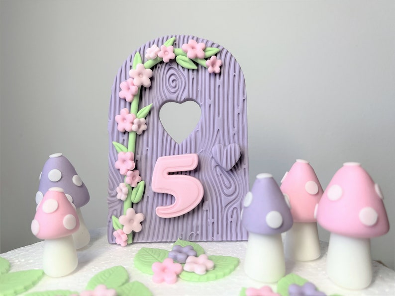 May include: Purple and pink fondant cake topper featuring a wooden door design, a heart-shaped window, and the number 5. The door is decorated with a vine of pink flowers and green leaves. There are also several purple and pink mushrooms with white polka dots around the door.