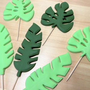 May include: Various green tropical leaf decorations on wooden sticks. The leaves are in different shades of green, from light to dark, and are arranged on a light brown surface. These are likely cake or cupcake toppers.