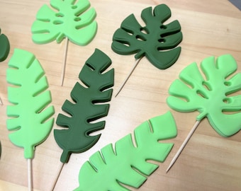 May include: An assortment of green tropical leaf-shaped decorations on wooden sticks. The leaves vary in shades of green, from light to dark, and are arranged on a light brown surface. These are likely cake or cupcake toppers.