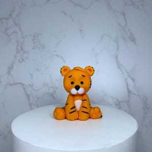 A small orange figurine of a tiger sitting on a white cake stand against a marble background.