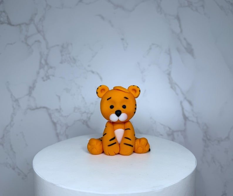 A small orange figurine of a tiger sitting on a white cake stand against a marble background.