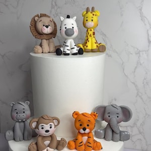 A collection of animal figurines, including a lion, zebra, giraffe, elephant, and monkey, are displayed on a white cake stand against a marble background.