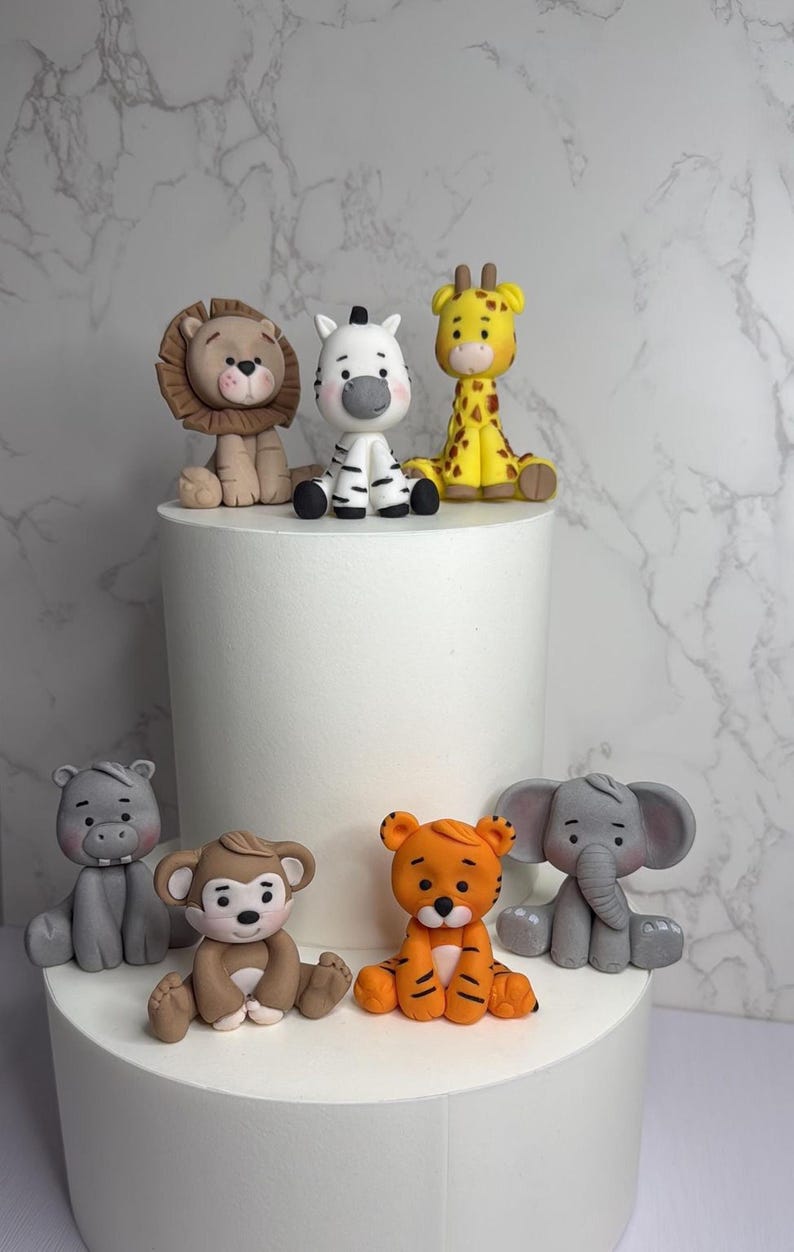 A collection of animal figurines, including a lion, zebra, giraffe, elephant, and monkey, are displayed on a white cake stand against a marble background.