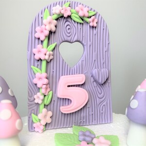 May include: Purple and pink fairy door cake topper with a heart-shaped window and a pink number 5. The door is decorated with a green vine and pink flowers. There are also purple and pink mushrooms with white polka dots around the door.