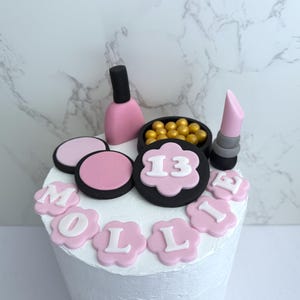 May include: A white cake decorated with pink and black fondant makeup-themed decorations. The decorations include a nail polish bottle, lipstick, blush, and a bowl of golden spheres. The cake is topped with the number 13 and the name "MOLLIE".
