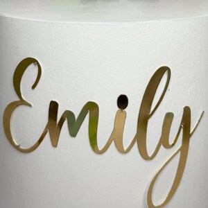 A white cylindrical object with the name "Emilly" written on it in gold cursive lettering.