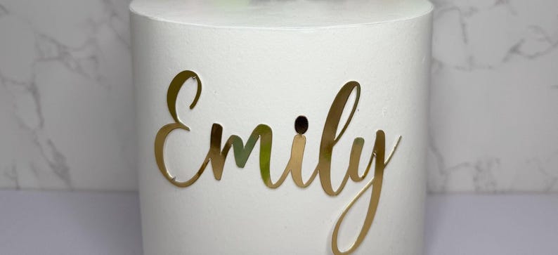A white cylindrical object with the name "Emilly" written on it in gold cursive lettering.