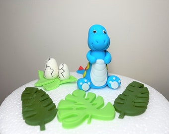 May include: Blue fondant dinosaur cake topper with white fondant eggs and green fondant leaves.