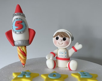 May include: A white fondant astronaut cake topper with a red and yellow rocket cake topper. The astronaut is wearing a white spacesuit with red accents and has brown hair. The rocket has a silver base and a red and yellow flame. The cake topper is decorated with the number 5 and the letters ROB.
