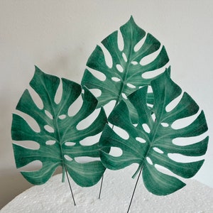 May include: Three green monstera leaves with cutouts, on black wire stems, arranged on a white cake.