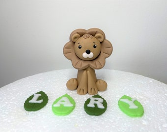 May include: A brown fondant lion cake topper with a white face and black eyes. The lion is sitting on a white cake with green fondant leaves spelling out the name "LARY".
