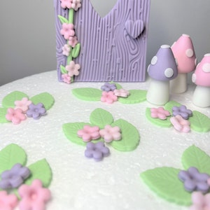 May include: A white cake decorated with purple and pink fondant flowers, green leaves, and purple and pink mushrooms.