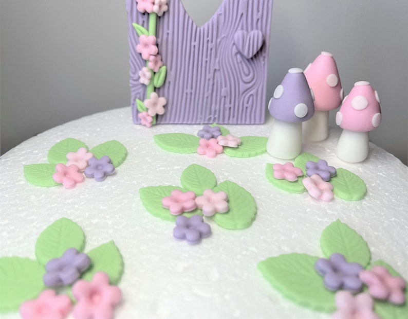 May include: A white cake decorated with purple and pink fondant flowers, green leaves, and purple and pink mushrooms.