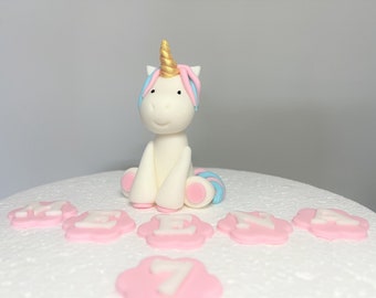 May include: A white fondant unicorn cake topper with a gold horn, blue and pink mane, and pink hooves. The unicorn is sitting on a white cake with pink fondant flowers and the number 7.