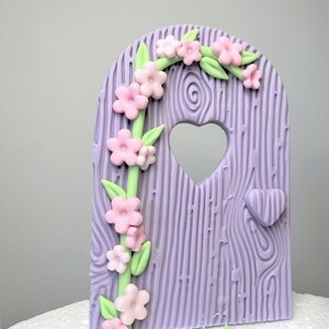 May include: A purple fondant cake topper shaped like a wooden door with a heart-shaped window and a vine of pink flowers and green leaves.