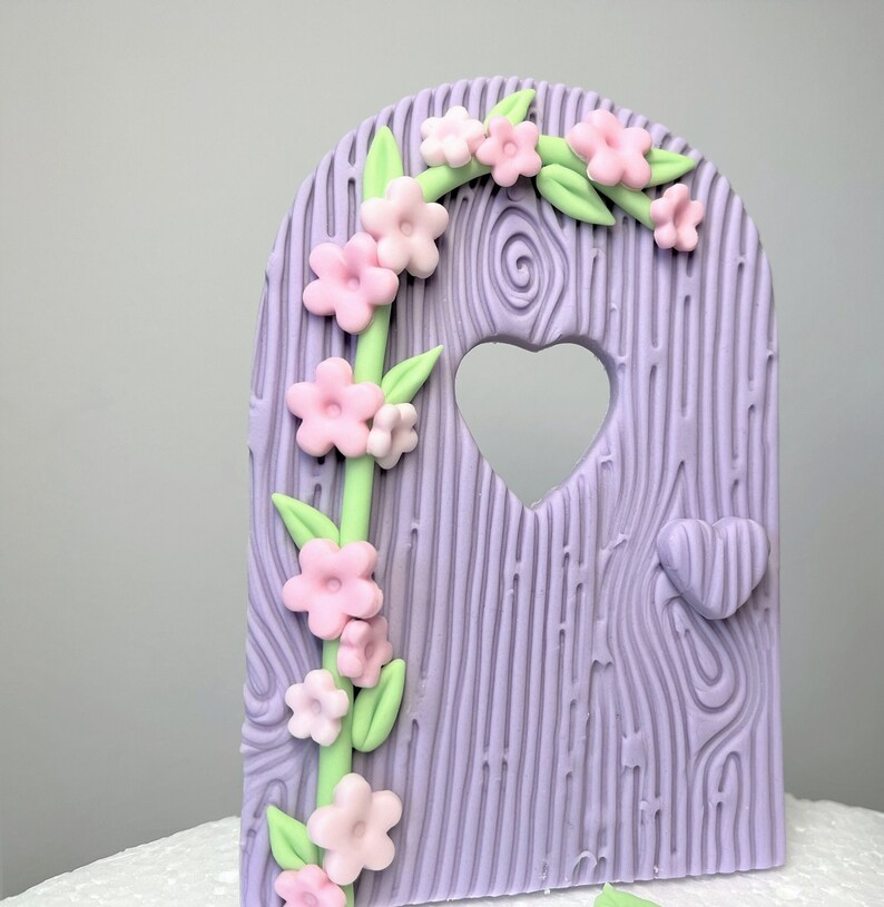 May include: A purple fondant cake topper shaped like a wooden door with a heart-shaped window and a vine of pink flowers and green leaves.