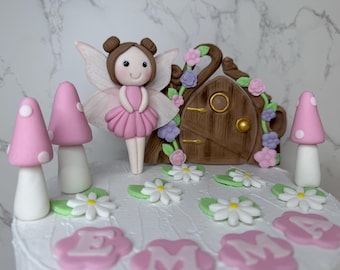May include: A cake decorated with a fairy, mushrooms, and a fairy house. The fairy has brown hair and wears a pink dress. Two pink mushrooms with white spots flank the fairy. The fairy house is brown with floral accents. White daisies and the name "EMMA" are also on the cake.