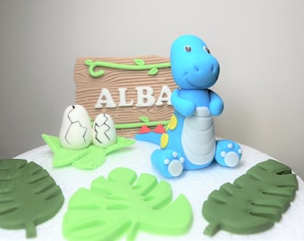 May include: A blue dinosaur cake topper with yellow spots and white feet, sitting on a white cake with green leaves and two white cracked eggs. A wooden sign with the name "ALBA" is in the background.