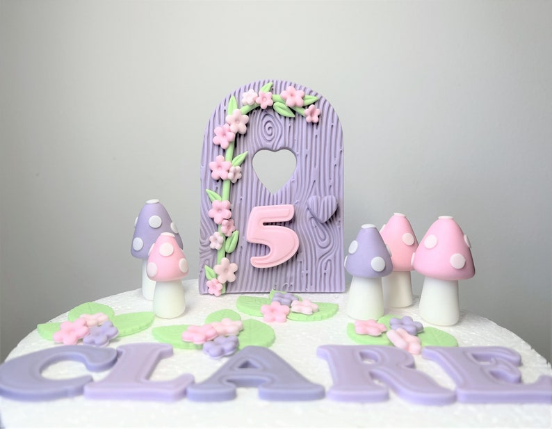 May include: Purple and pink fairy door cake topper with a heart-shaped window and the number 5. The topper is surrounded by pink and green flowers and mushrooms. The name "CLAIRE" is written in purple icing on the cake.