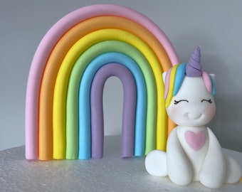 May include: A white unicorn figurine with a rainbow mane and a pink heart on its chest, sitting in front of a pastel rainbow cake topper.