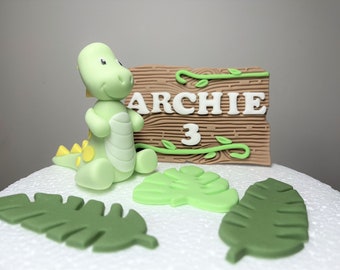 May include: A green dinosaur cake topper with a wooden sign that says "Archie 3" and green leaves.