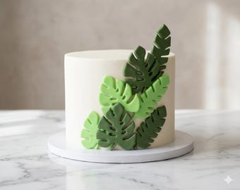 May include: A cylindrical cake with smooth white frosting, decorated on the side with an arrangement of layered, cut-out monstera leaves in varying shades of light and dark green, resting on a circular white base.