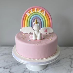 A pink birthday cake decorated with a unicorn and a rainbow, with the name "ELIZA" written on the cake.