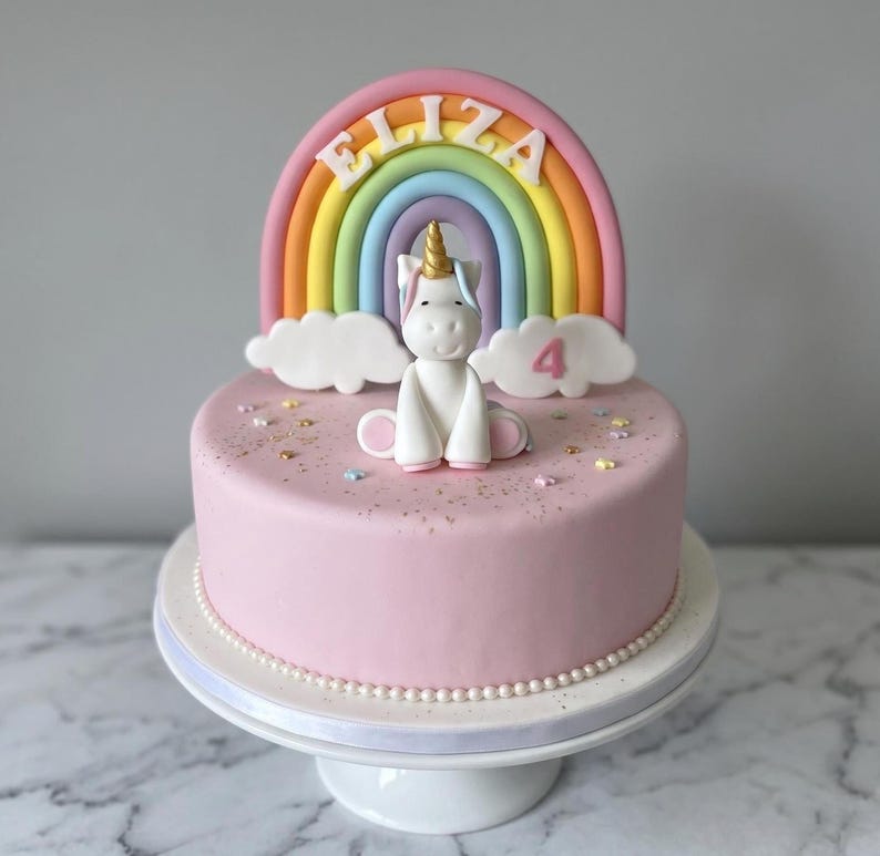 A pink birthday cake decorated with a unicorn and a rainbow, with the name "ELIZA" written on the cake.