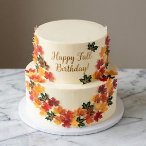 Può includere: Una torta a due piani con glassa color avorio, decorata con foglie autunnali colorate. La torta presenta la scritta dorata "Happy Fall Birthday!". Poggia su un vassoio bianco.