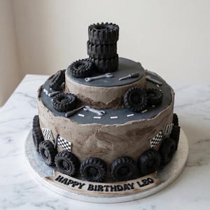 Puede incluir: Tarta de dos pisos decorada con tem&aacute;tica de coches de carreras. La tarta presenta neum&aacute;ticos negros, herramientas y banderas a cuadros. El piso superior tiene una pila de neum&aacute;ticos. El texto "HAPPY BIRTHDAY LEO" es visible.