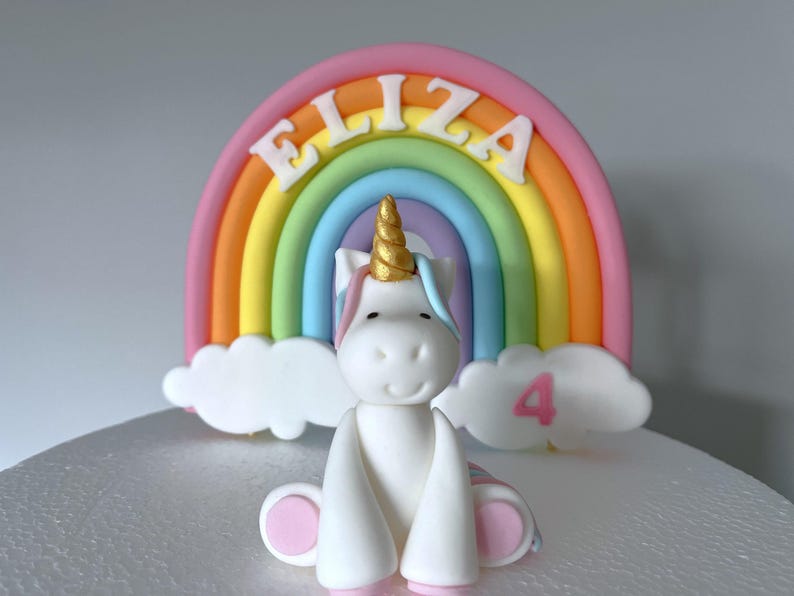 A white unicorn figurine with a golden horn sits in front of a rainbow-colored cake topper with the name "Eliza" written on it.