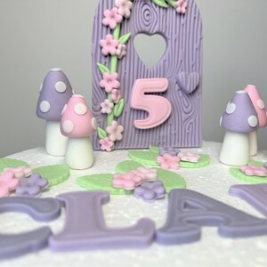 May include: A purple and pink fairy garden themed cake with the name "CLARA" and the number "5" on it. The cake is decorated with mushrooms, flowers, and leaves.