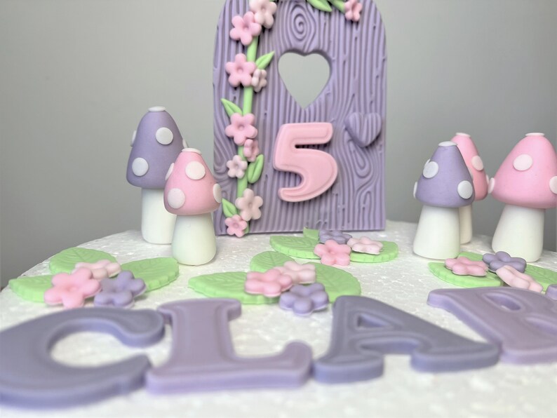 May include: A purple and pink fairy garden themed cake with the name "CLARA" and the number "5" on it. The cake is decorated with mushrooms, flowers, and leaves.